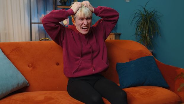 Scared woman with short blonde hair screams from stress tension problems panic horror hopelessness fear surprise shock expresses gestures rage. Furious girl at home in room on orange couch. Lifestyle