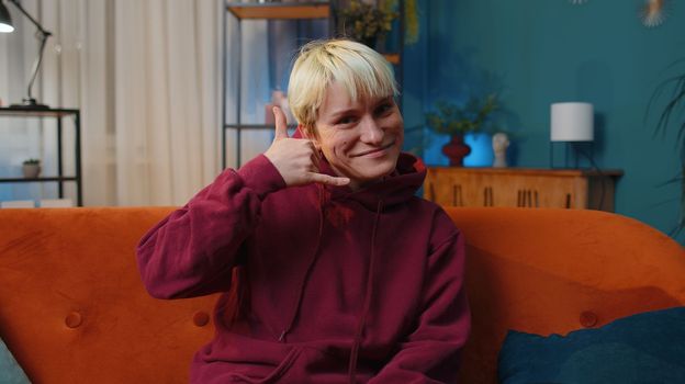 Call me, here is contact number. Young woman with short blonde hair looking at camera advertising doing phone gesture like says hey you call me back at home. Lovely girl at evening room apartment