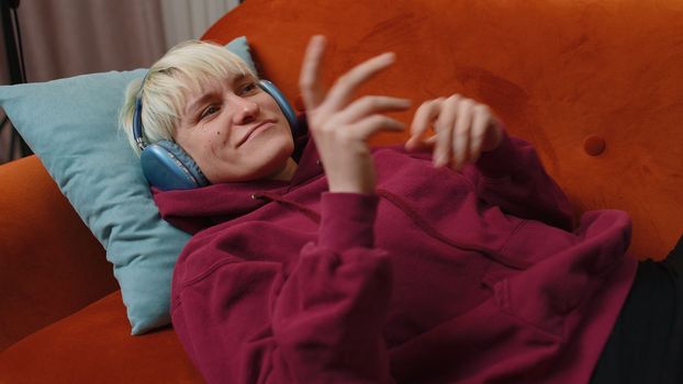 Happy overjoyed woman with short hair in wireless headphones relaxing sitting on couch at home choosing listening favorite energetic disco dancing music. People weekend evening leisure activities