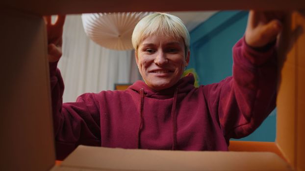 Happy surprised winner woman unpacking delivery parcel at home. Smiling satisfied girl shopper online shop customer opening cardboard box receiving purchase gift headphones by fast postal shipping