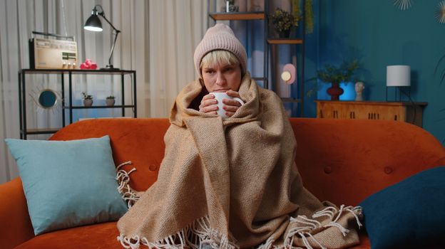 Sick young woman wrapped in plaid sitting, shivering from cold on sofa drinking hot tea in unheated apartment without heating due debt. Unhealthy girl feeling discomfort try to warm up at evening home