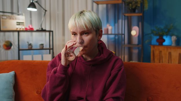 Thirsty woman with short blonde hair holding glass of natural aqua make sips drinking still water preventing dehydration at home. Girl with good life habits, healthy slimming, weight loss concept