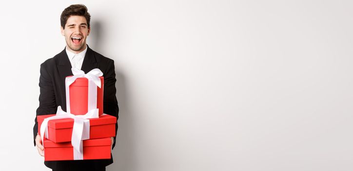 Image of handsome guy in black suit, holding gifts and winking at camera, have christmas presents, standing against white background.