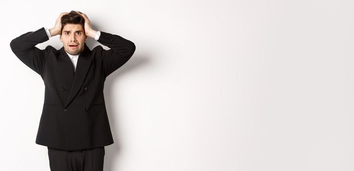 Frustrated and worried businessman in black suit, panicking as looking at trouble, holding hands on head alarmed, standing against white background.