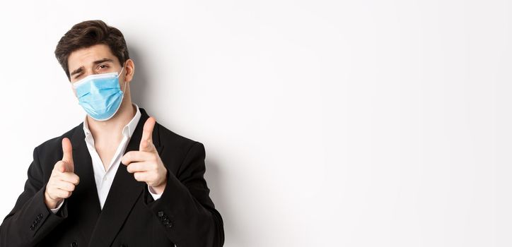 Concept of covid-19, business and social distancing. Close-up of sassy man in trendy suit and medical mask, pointing fingers at camera, standing against white background.