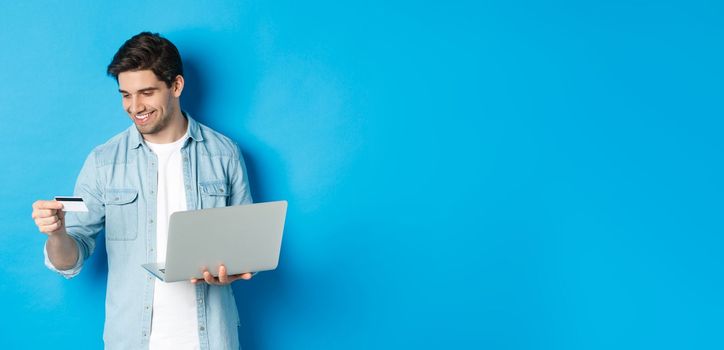 Young man order online, holding credit card and laptop, shopping in internet, standing against blue background.