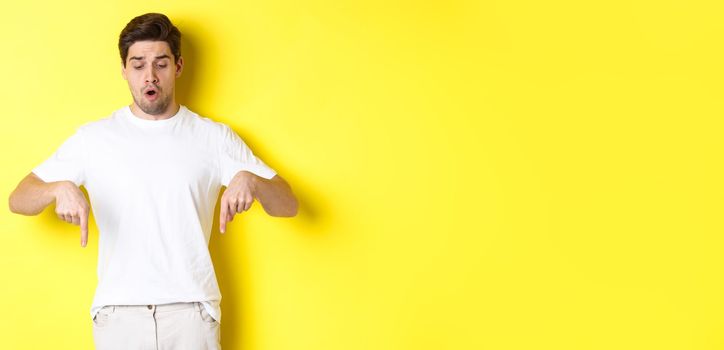 Worried guy pointing fingers and looking down, standing startled against yellow background.