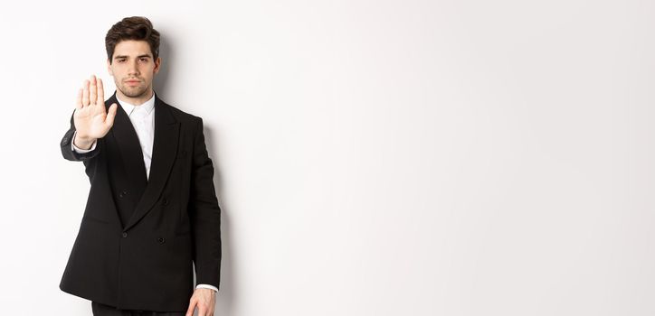 Portrait of serious handsome man in formal suit, extending hand to stop you, prohibit action, forbid and disagree with something, standing against white background.