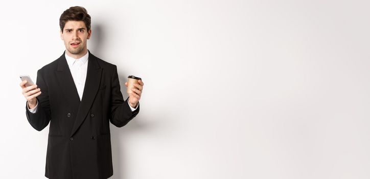 Image of confused businessman standing in a suit, holding cup of coffee and mobile phone, cant understand something, standing against white background.