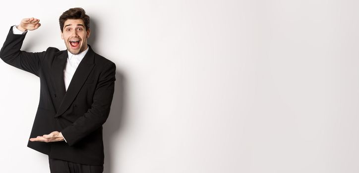 Portrait of excited handsome man in suit, shaping big object on copy space and smiling amazed, holding something, standing over white background.