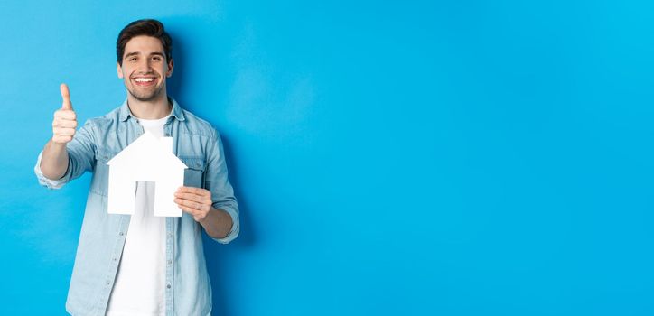 Insurance, mortgage and real estate concept. Satisfied client showing house model and thumb up, smiling pleased, standing against blue background.