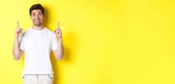Excited attractive man in white t-shirt, pointing fingers up, looking at advertisement with happy smile, standing over yellow background.