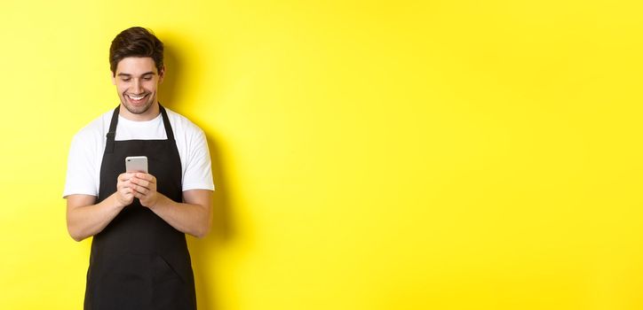 Waiter in black apron reading message on mobile phone, smiling happy, standing over yellow background.