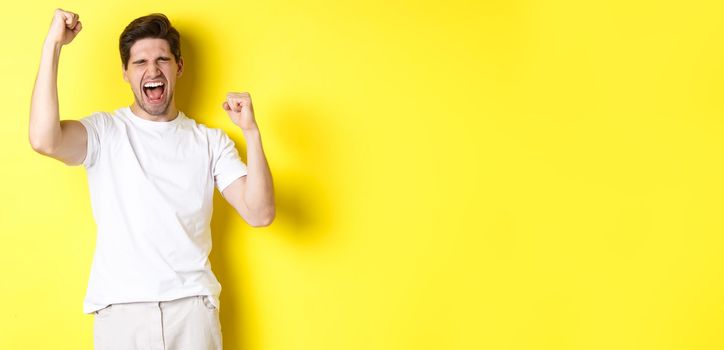 Excited handsome man winning and shouting for joy, raising hand up and triumping, achieve goal, standing over yellow background.