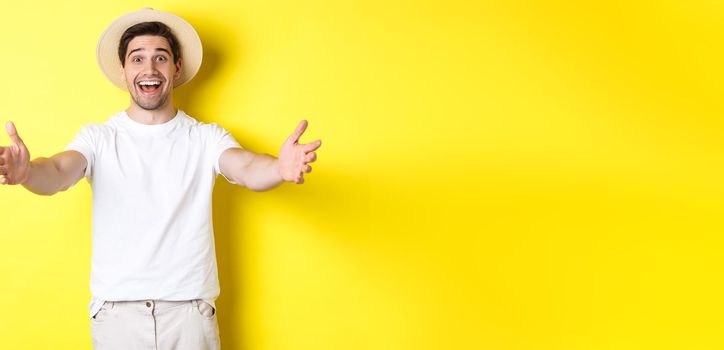 Concept of tourism and summer. Happy and friendly man traveller reaching hands for hug, greeting or welcome you, standing over yellow background.