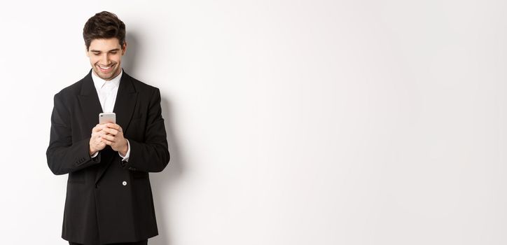 Portrait of handsome stylish businessman in black suit, writing a message, smiling and looking at smartphone, standing over white background.