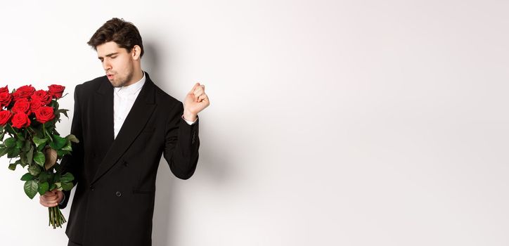 Image of elegant and sassy man in black suit, looking confident and holding bouquet of red roses, going on a romantic date, standing against white background.