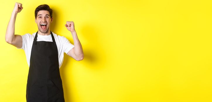 Happy barista celebrating victory, raising hands up and shouting for joy, wearing black apron, shop uniform, standing against yellow background.