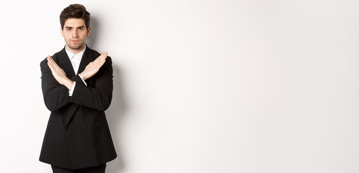 Portrait of serious and concerned man in black suit, showing stop gesture and frowning, making cross to prohibit or decline something, standing over white background.