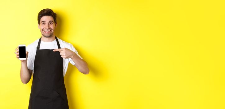 Handsome barista in black apron pointing finger at mobile screen, showing app and smiling, standing over yellow background.