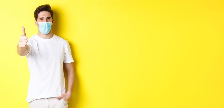 Concept of coronavirus, pandemic and social distancing. Confident young man in medical mask showing thumbs up and winking, yellow background.