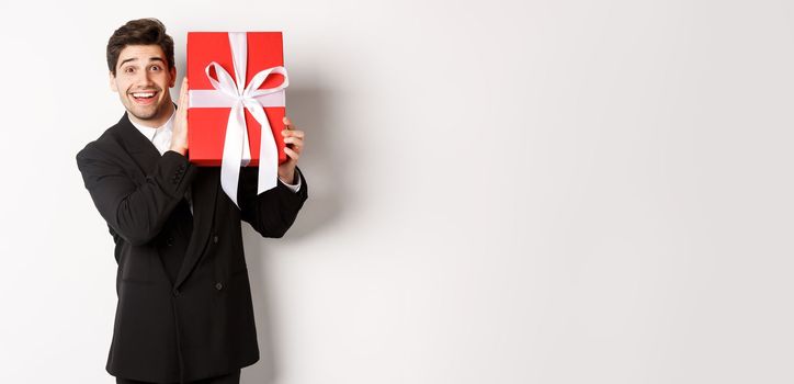 Handsome man in black suit, receiving christmas gift, smiling amazed, standing against white background.