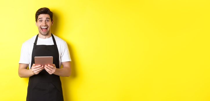 Happy salesman in black apron, holding digital tablet and looking surprised, standing against yellow background.