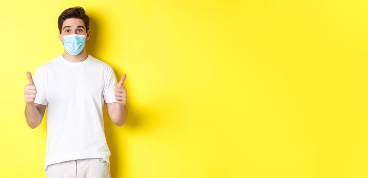 Concept of covid-19, quarantine and lifestyle. Happy man in medical mask showing thumbs up, approve or saying yes, like something good, yellow background.