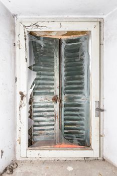 Broken window. Behind, a gray roller shutter that protect the house from the draft of air. Abandonment.