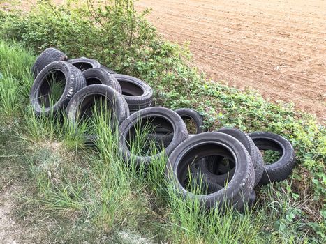 A pile of old car tires abandoned on the grass near a field. A dump of old used car tires. Environmental pollution.