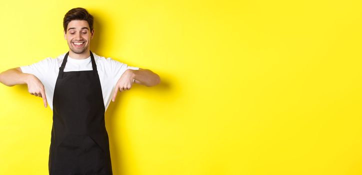 Happy coffee shop worker in black apron pointing fingers down, looking pleased and smiling, showing logo, standing over yellow background.