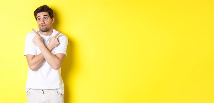 Indecisive man shrugging, pointing fingers sideways, troubled to choose, standing over yellow background.