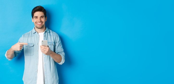 Concept of online shopping, applications and technology. Handsome man recommending app on smartphone, pointing at phone and smiling satisfied, standing over blue background.