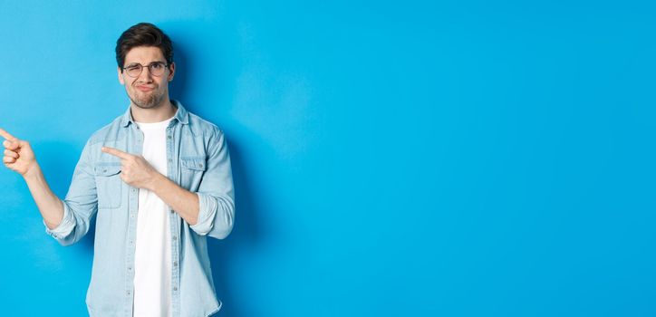 Displeased and skeptical male model in glasses, pointing fingers left at something bad, showing awful advertisement, standing over blue background.