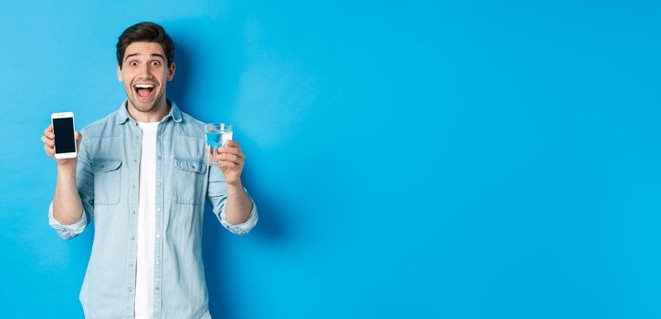 Happy man showing glass of water and mobile screen, recommending smartphone health app, standing over blue background.