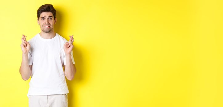 Nervous guy crossing fingers for good luck, hoping for something, standing over yellow background.