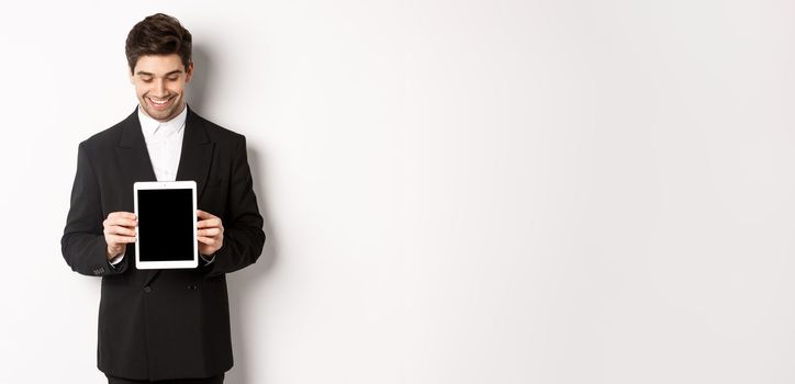 Image of good-looking male entrepreneur in black suit, looking down at digital tablet screen and showing advertisement, standing against white background.