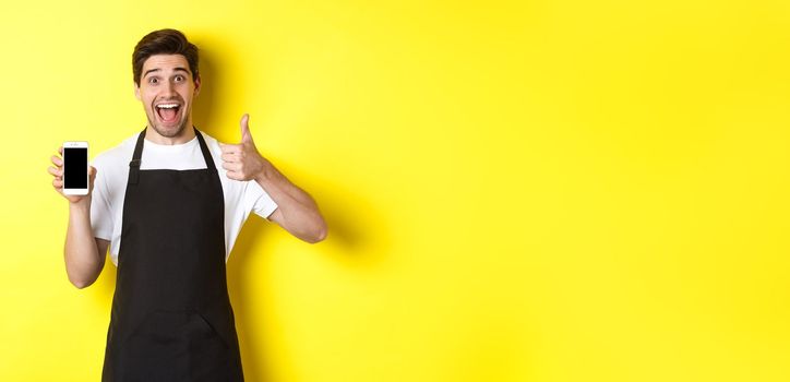 Happy barista in black apron showing smartphone screen, make thumb-up, recommending cafe application, standing over yellow background.