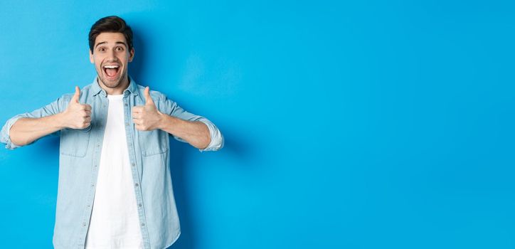 Smiling adult man showing thumbs up with excited face, like something awesome, approving product, standing against blue background.