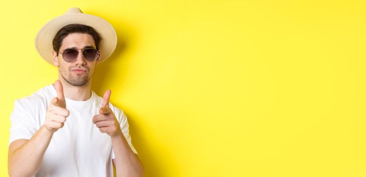 Concept of tourism and vacation. Close-up of cool guy in summer hat and sunglasses pointing finger guns at camera, standing over yellow background.
