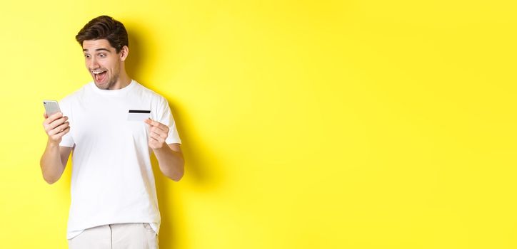 Man looking surprised at smartphone, shopping online, holding credit card, standing over yellow background. Copy space