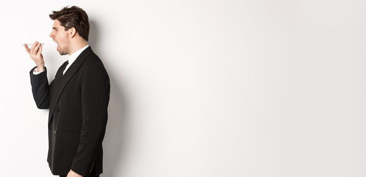 Profile shot of angry businessman in black suit, shouting at speakerphone and looking mad, recording voice message, standing over white background.