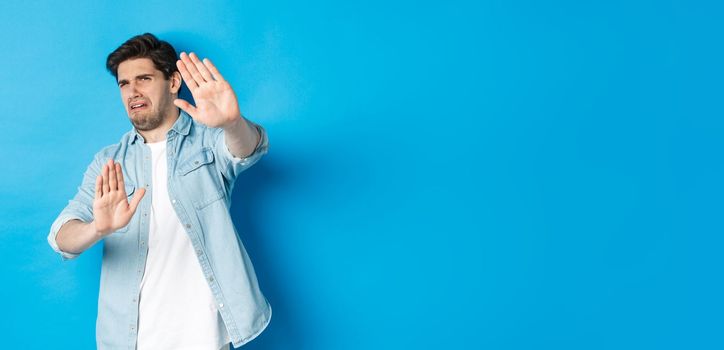 Disgusted guy saying no, refusing and looking away from something awful, cringe while standing against blue background.