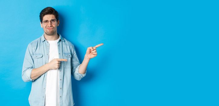 Disappointed young man in glasses pointing fingers right at copy space, showing promo banner and smirking dissatisfied, judging bad product, standing over blue background.