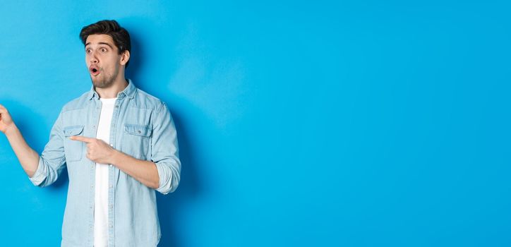 Surprised and amazed man looking at promotion, pointing fingers left at advertisement, standing over blue background.