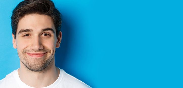 Headshot of attractive man smiling pleased, looking intrigued, standing over blue background.