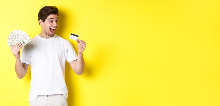 Cheerful guy looking at credit card, holding money, concept of bank credit and loans, standing over yellow background.