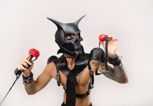 A sexy man in bdsm wearing a demon mask with a whip wearing a leather cloak with leather handcuffs and partuppets on a white background