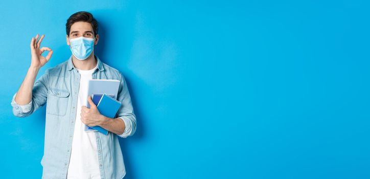 Education, covid-19 and social distancing. Guy student in medical mask looking happy, holding notebooks, showing ok sign, standing over blue background.