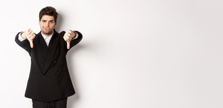 Portrait of skeptical and disappointed man in black suit, frowning upset, showing thumbs-down, dislike something bad, standing over white background.